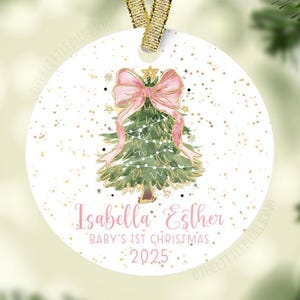 May include: A white Christmas ornament with a watercolor illustration of a Christmas tree adorned with a pink bow and lights. The ornament has the text "Isabella Esther, Baby's 1st Christmas, 2025" in pink script. A gold ribbon is attached for hanging.