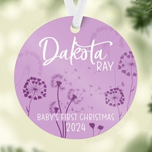 May include: A round, lavender Christmas ornament with the name "Dakota Ray" in white script. The ornament features a dandelion design and the text "Baby's First Christmas 2024". A white ribbon is attached for hanging.