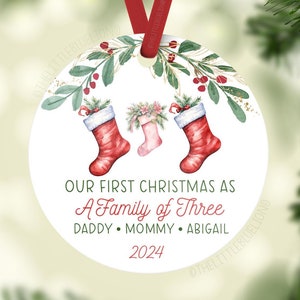 May include: A white round Christmas ornament with a watercolor illustration of two red stockings with white trim and a small stocking in the middle. The text reads "Our First Christmas as A Family of Three Daddy Mommy Abigail 2024".