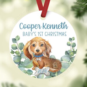 May include: A white round Christmas ornament with a watercolor illustration of a brown dachshund puppy wearing a blue collar. The ornament has the text "Cooper Kenneth Baby's 1st Christmas 2024" on it.