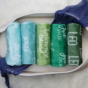 May include: A set of six personalized baby blankets rolled up and arranged in a white wicker basket. The blankets are in shades of blue and green with white lettering. The names on the blankets are Zaedyn, Ronan, Lawrence, Edward, Jack Dylan, and Leo.