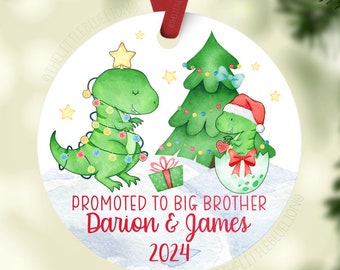 May include: A white round Christmas ornament with a green dinosaur wearing Christmas lights, a green Christmas tree, and a dinosaur in an egg wearing a Santa hat. The ornament says "Promoted to Big Brother Darian & James 2024".
