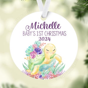 May include: A white, round Christmas ornament with a watercolor illustration of a turtle and succulents. The ornament reads "Michelle Baby's 1st Christmas 2024" in purple script. A white ribbon is attached for hanging.
