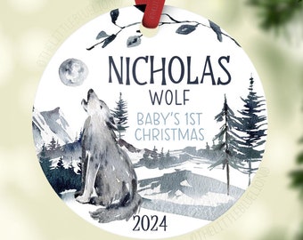 May include: A round Christmas ornament with a watercolor design featuring a howling wolf, mountains, and pine trees. The ornament has the text "NICHOLAS WOLF BABY'S 1ST CHRISTMAS 2024" and is suspended by a red ribbon.