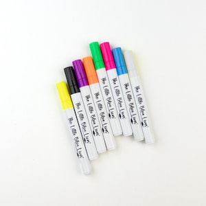 May include: Eight white dry-erase markers with different colored tips. The markers are arranged in a fan shape with the text "The Little Blue Lion" printed on each marker.