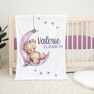 Puede incluir: Una manta de beb&eacute; blanca con una luna creciente morada y un oso de peluche durmiendo sobre ella. La luna tiene un dise&ntilde;o floral y el texto "Valerie ELIZABETH" est&aacute; impreso en la manta.