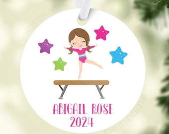 Op de afbeelding: Een witte ronde kerstbal met een cartoonillustratie van een meisje-turnster op een evenwichtsbalk. Het meisje draagt een roze turnpakje met een regenboogstreep en heeft bruin haar. Er zijn vier sterren om haar heen, in roze, paars, blauw en groen. De kerstbal heeft ook de tekst "Abigail Rose 2024" in roze.
