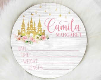 Könnte beinhalten: Ein rundes Schild aus weißem Holz mit einem rosafarbenen und goldenen Schlossdesign. Das Schild trägt den Namen "Camila Margaret" in rosa und goldenen Buchstaben. Das Schild hat auch Platz zum Schreiben von Datum, Uhrzeit, Gewicht und Länge.