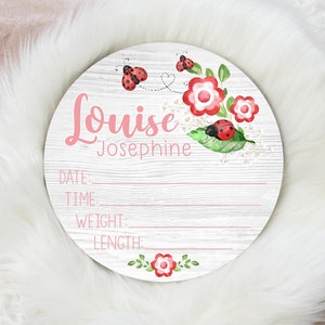 May include: A white wooden round sign with a pink and white floral design and ladybugs. The sign says "Louise Josephine" and has spaces to fill in the date, time, weight, and length.