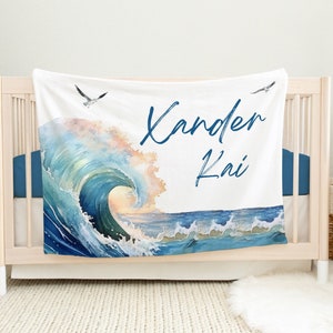 May include: White baby blanket with a watercolor painting of a blue ocean wave and seagulls. The blanket has the names "Xander Kai" written in blue cursive.