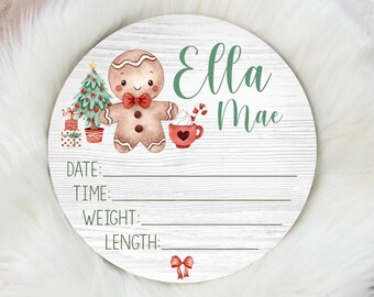 May include: A round wooden sign with a gingerbread man, a Christmas tree, and a cup of hot chocolate. The sign has the name "Ella Mae" written on it and spaces to fill in the date, time, weight, and length.