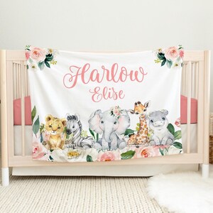 K&ouml;nnte beinhalten: Eine wei&szlig;e Babydecke mit einem rosafarbenen Blumendesign und einem rosafarbenen Namen "Harlow Elise" in Kursivschrift. Die Decke zeigt eine Reihe von niedlichen Tierillustrationen, darunter ein L&ouml;we, ein Zebra, ein Elefant, eine Giraffe und ein Flusspferd.