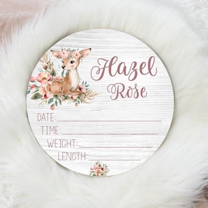 May include: A round wooden sign with a whitewashed background and a pink floral design. A watercolor illustration of a fawn with a floral crown is in the upper left corner. The sign says "Hazel Rose" in pink script and has blank lines for "Date", "Time", "Weight", and "Length".