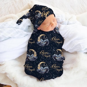 May include: A newborn baby swaddled in a navy blue blanket and matching cap, both adorned with a celestial pattern of gold moons, stars, and clouds. The name "Raven" is printed in gold script on the cap and blanket.