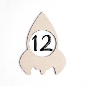 May include: Wooden rocket-shaped milestone marker with a circle cutout for a number. The number 12 is written in black marker inside the circle. The marker is made of 1/4 inch Baltic birch wood and is sanded and ready to paint.