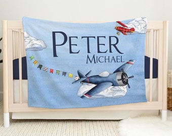 Puede incluir: Una manta para bebé azul claro con un diseño de acuarela con aviones, nubes y una pancarta de banderas de colores. El nombre "PETER MICHAEL" está impreso en azul marino. La manta está sobre una cuna.