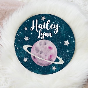 May include: A teal blue round sign with a pink planet and silver glitter stars. The sign says "Hailey Lynn" in white cursive lettering.