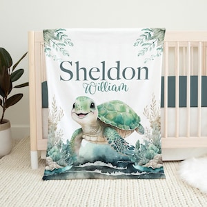 May include: A white baby blanket with a green sea turtle illustration. The turtle is swimming in blue water with green seaweed and coral. The blanket has the names "Sheldon William" printed in gray text.
