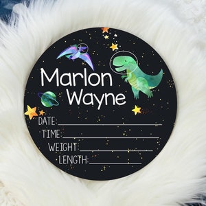 May include: Black and white round wooden sign with a space theme. It features a green dinosaur, a purple and blue pterodactyl, and stars. The sign says "Marlon Wayne" and has spaces to fill in the date, time, weight, and length.