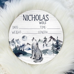 May include: A white wooden round sign with a watercolor illustration of a wolf howling at the moon in a snowy mountain landscape. The sign says "Nicholas Wolf" and has spaces to fill in the date, time, weight, and length.