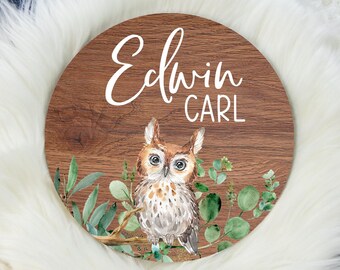May include: A round wooden sign with a brown woodgrain finish. The sign features a watercolour illustration of an owl perched on a branch with green leaves. The text "Edwin Carl" is written in white cursive lettering on the sign.