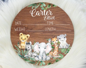 May include: A round wooden sign with a wood grain pattern. The sign features the names "Carter Elliot" in script. Below the names are spaces for date, time, weight, and length. The bottom of the sign has illustrations of a lion, zebra, elephant, giraffe, and hippo.