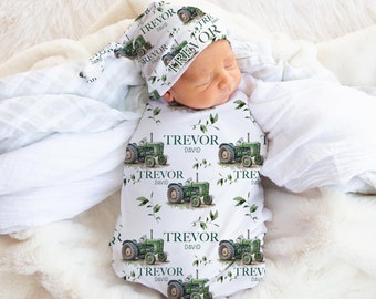 May include: A white baby onesie with a green tractor print and the name "Trevor David" repeated throughout. The onesie is paired with a matching green and white hat with the name "Trevor" on it. The baby is sleeping on a white blanket.