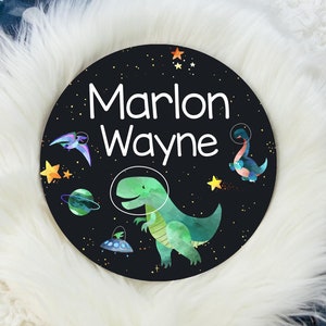 May include: A black and white round sign with a space theme. The sign has a green dinosaur, a spaceship, stars, and planets. The text "Marlon Wayne" is written in white on the sign.