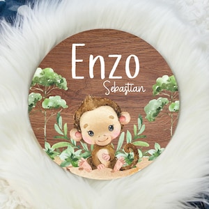 May include: A round wooden sign with a brown woodgrain background. The sign features a cartoon monkey sitting in a jungle scene with green leaves. The sign is personalized with the name "Enzo Sebastian".