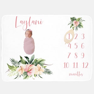 May include: A white baby milestone blanket with a floral design and the name "Laylani" in pink script. The blanket features a baby wrapped in a pink blanket, a wooden cutout with the number 1, and numbers 1-12 for monthly milestones.