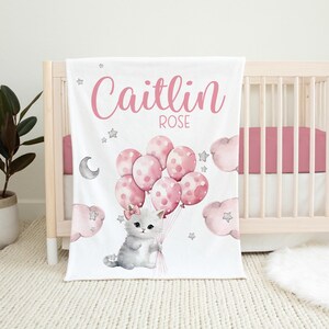 May include: A white baby blanket with a pink and white cat holding pink balloons with white polka dots. The blanket has the name "Caitlin Rose" written in pink cursive.