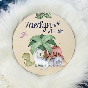K&ouml;nnte beinhalten: Ein rundes Holzschild mit Dinosaurier-Motiv. Das Schild zeigt einen gr&uuml;nen Palmenbaum, einen Vulkan und vier Dinosaurier. Der Text "Zaedyn William" ist in Lila auf dem Schild geschrieben.