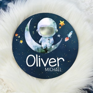 May include: A blue and white round sign with a crescent moon, stars, and a cartoon astronaut. The sign says "Oliver Michael" in white text.