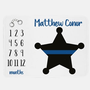 May include: White baby milestone blanket with a black and blue police badge design. The blanket has the numbers 1-12 and the words "months" printed on it. The name "Matthew Conor" is printed at the top of the blanket.
