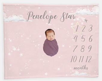 May include: Pink baby milestone blanket with white stars and clouds. The blanket features a gold star with numbers 1-12 for tracking a baby's growth. The text "Penelope Star" is written in gray cursive. A baby is pictured in the center of the blanket.