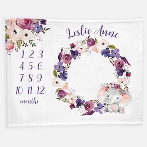 May include: A white baby blanket with a purple floral wreath and a gray elephant with a pink heart. The blanket has a number chart for tracking a baby's growth and the name "Leslie Anne" is written at the top.