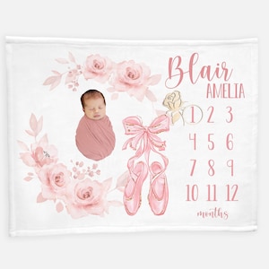 May include: Pink and white baby milestone blanket with a floral design, a baby photo, and pink ballet shoes. The blanket has the text "Blair Amelia" and numbers 1-12 for tracking baby's growth.