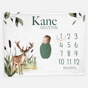 May include: A white baby blanket with a green deer and a baby boy in a green swaddle. The blanket has the name "Kane Houston" and a number chart for tracking a baby's growth from 1 to 12 months.