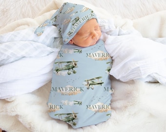 May include: A baby swaddled in a blue and white swaddle with a repeating pattern of airplanes and the name "Maverick Wright". The baby is wearing a matching blue and white hat with the name "Maverick" on it.