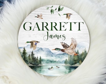 May include: A round wooden sign with a watercolor scene of a lake, mountains, and flying ducks. The sign has the name "GARRETT James" written in green text.