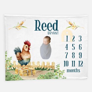 May include: A white baby blanket with a watercolor illustration of a rooster, a fence, and a barn. The blanket also features the name "Reed Bryant" and a chart with numbers 1 through 12 for tracking a baby's monthly growth.
