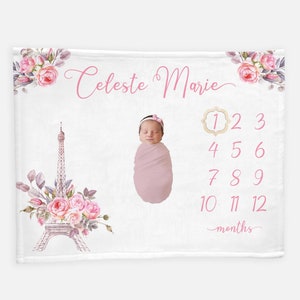 May include: A white baby milestone blanket with a pink floral design, a pink Eiffel Tower, and a pink baby girl wrapped in a pink blanket. The blanket has a number chart for each month of the baby's first year. The text "Celeste Marie" is written in pink cursive at the top of the blanket.