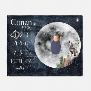 May include: A navy blue baby milestone blanket with a watercolor illustration of a wolf howling at the moon. The blanket features the name "Conan Wolfe" and numbers for tracking baby's age in months.