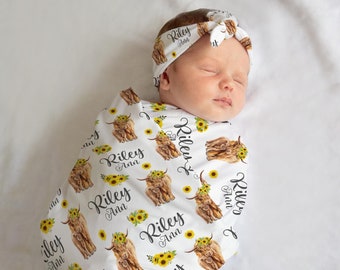 May include: A white swaddle blanket with a brown and white Highland cow pattern and yellow sunflowers. The blanket has the name "Riley Ann" repeated throughout the design. A baby is wrapped in the blanket and wearing a matching headband.