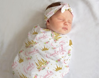 Könnte beinhalten: Eine weiße Wickeldecke mit einem rosa und goldenen Schloss- und Kronenmuster. Die Decke trägt den Namen "Camila Margaret", der sich über das gesamte Muster wiederholt. Ein Baby schläft in der Wickeldecke und trägt ein rosa und goldenes Stirnband mit dem Namen "Camila" darauf.