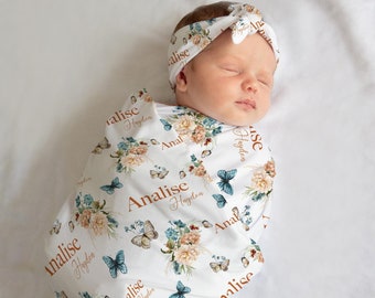 May include: A white swaddle blanket with a floral and butterfly pattern. The blanket has the name "Analise Hayden" repeated throughout the design. A baby is swaddled in the blanket and is wearing a matching headband with the same name.