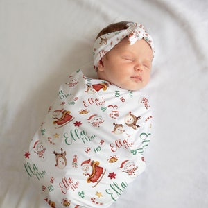 K&ouml;nnte beinhalten: Eine wei&szlig;e Wickeldecke mit Weihnachtsmotiven wie Weihnachtsmann, Rentier, Schneemann und Zuckerstangen. Die Decke tr&auml;gt den Namen "Ella Mae", der sich &uuml;ber das gesamte Design wiederholt. Ein Baby ist in die Decke gewickelt und tr&auml;gt ein passendes Stirnband.