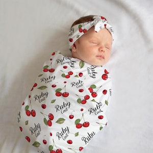 K&ouml;nnte beinhalten: Eine wei&szlig;e Swaddle-Decke mit einem roten Kirschmuster und dem Text "Ruby Cait", der sich wiederholt. Ein Baby ist in die Decke gewickelt und tr&auml;gt ein passendes Stirnband.