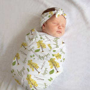 May include: A white swaddle blanket with a repeating pattern of yellow and green birch trees and the name "Aspyn" written in green. A baby is sleeping inside the swaddle, wearing a white headband with the same pattern as the swaddle.