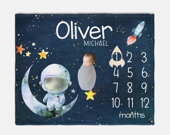 May include: A blue baby blanket with a space theme. It features a cartoon astronaut standing on a crescent moon, a rocket ship, and stars. The blanket also has a number chart for tracking a baby's growth, with the name "Oliver Michael" printed at the top.